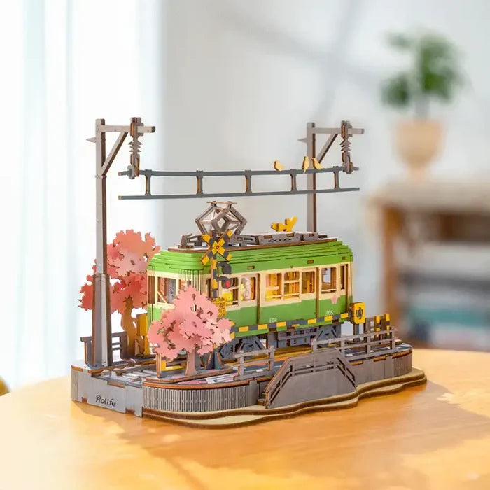 Modèle décoratif d’un trolley vert à l’arrêt d’une gare avec des cerisiers roses en fleurs et des lignes électriques aériennes.